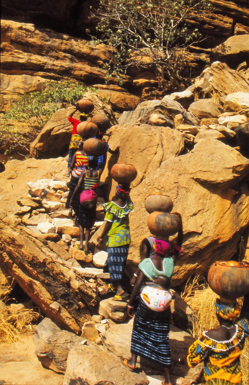 Dogon women climbing a rock slope carrying pots on their heads on the path to market in Mali.