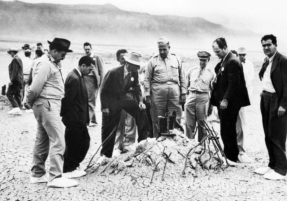 J. Robert Oppenheimer in a light-colored hat with his foot on rubble, standing with General Leslie Groves and others at the Trinity test ground zero