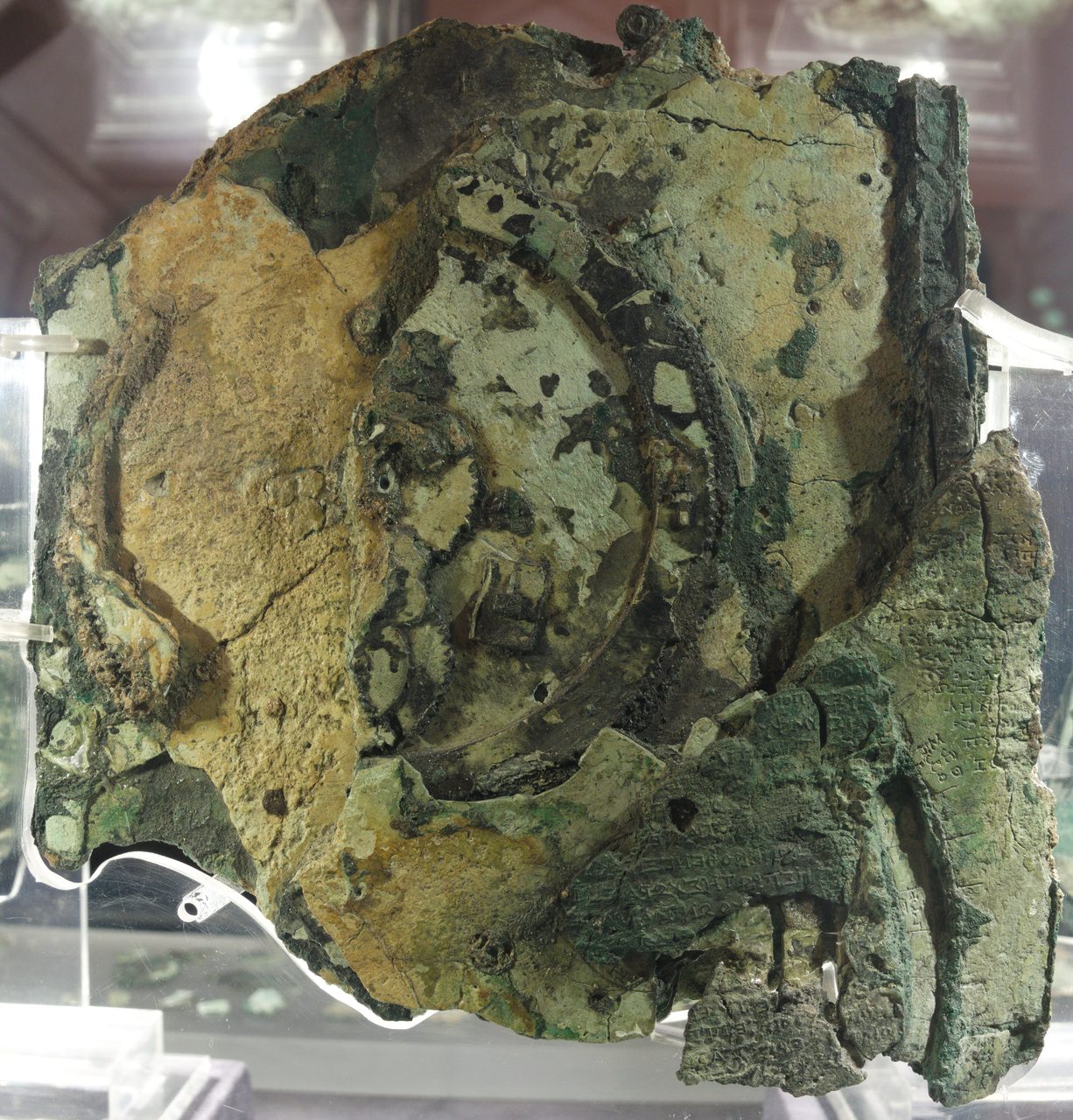 The rear face of Fragment A of the Antikythera Mechanism, a corroded block of bronze wheels and plates with incised Greek lettering visible on the plate at right