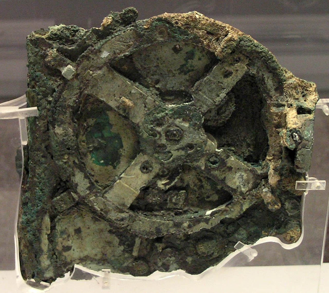 Close-up of Fragment A of the Antikythera Mechanism, a corroded green bronze block with a large spoked gear wheel on its surface, held on clear mounts in a museum case