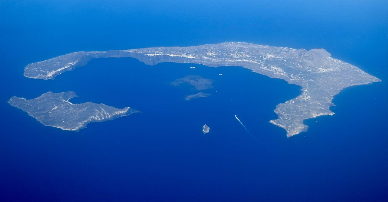 An aerial view of the flooded volcanic caldera of Santorini, with white cliff-top buildings along the rim
