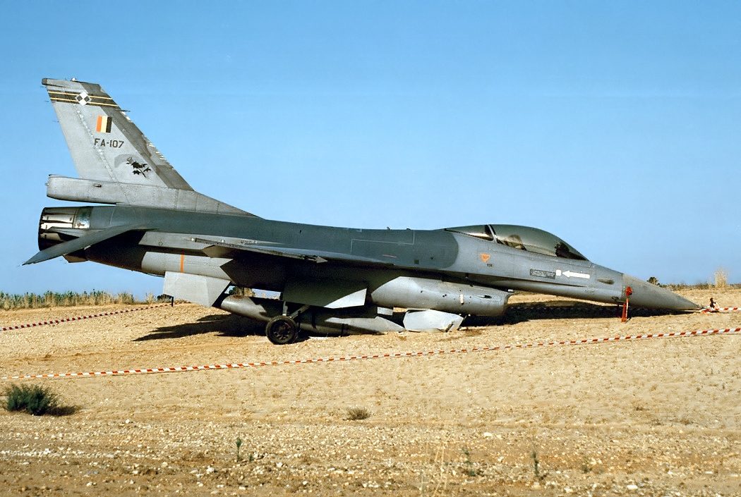 A Belgian Air Force F-16A fighter on bare ground behind red and white barrier tape, with FA-107 painted on its fin