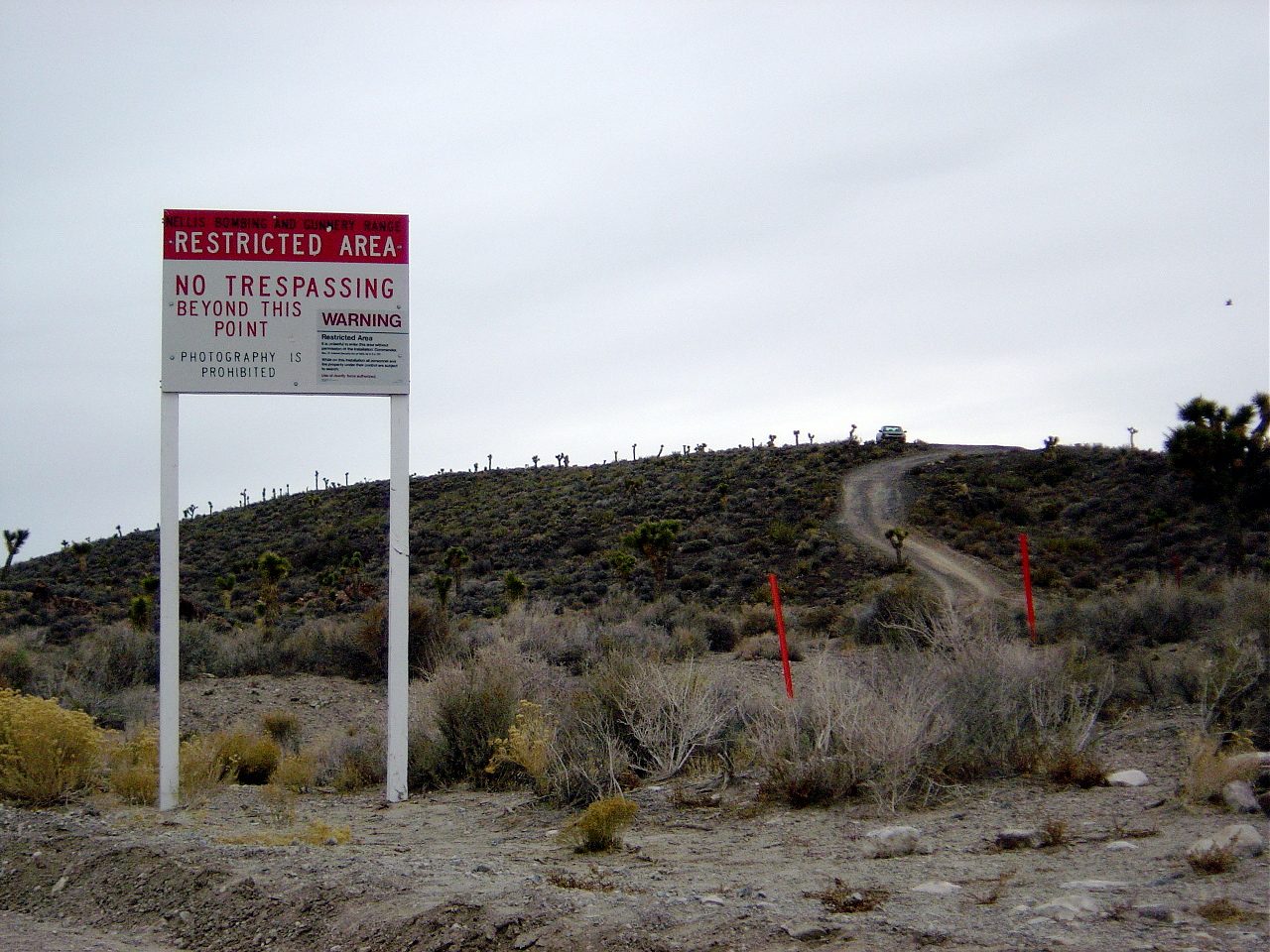 A warning sign in the Nevada desert reading NELLIS BOMBING AND GUNNERY RANGE, RESTRICTED AREA, NO TRESPASSING BEYOND THIS POINT and PHOTOGRAPHY IS PROHIBITED, with a smaller warning panel set into it.