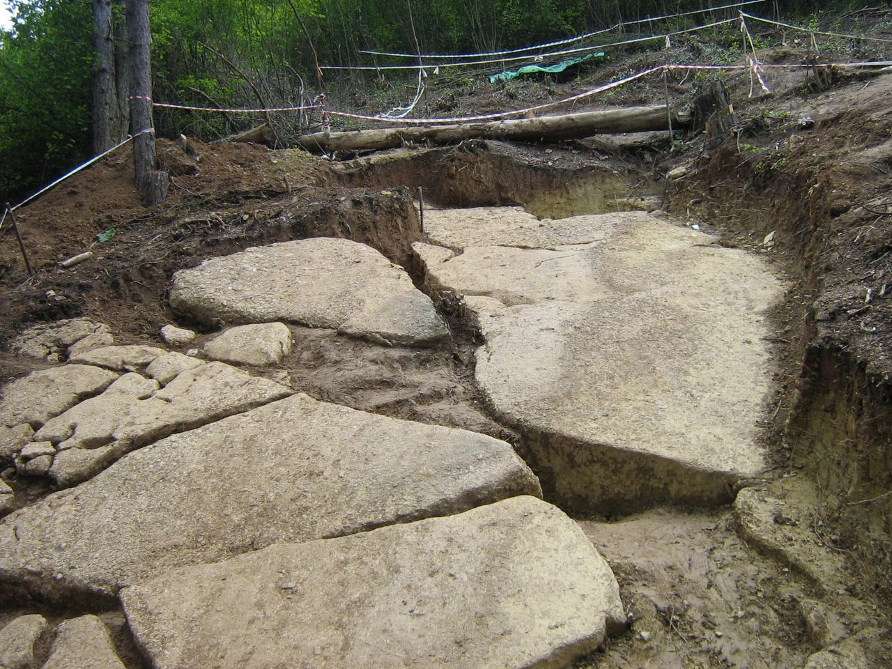 An excavation trench cut into the hillside at Visočica, exposing a broad pavement of large pale flat-topped stone slabs separated by straight joint lines, with cut earth banks, felled logs and red-and-white barrier tape behind