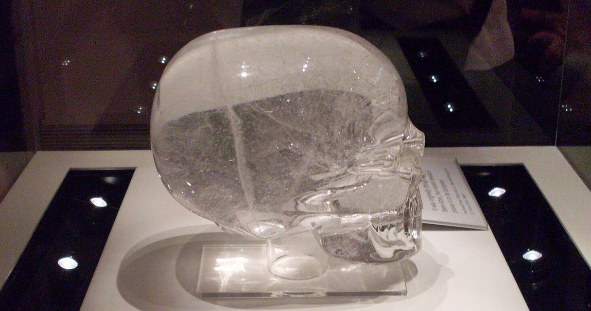 A life-size human skull carved from clear rock crystal, photographed in a museum case on a pale ground