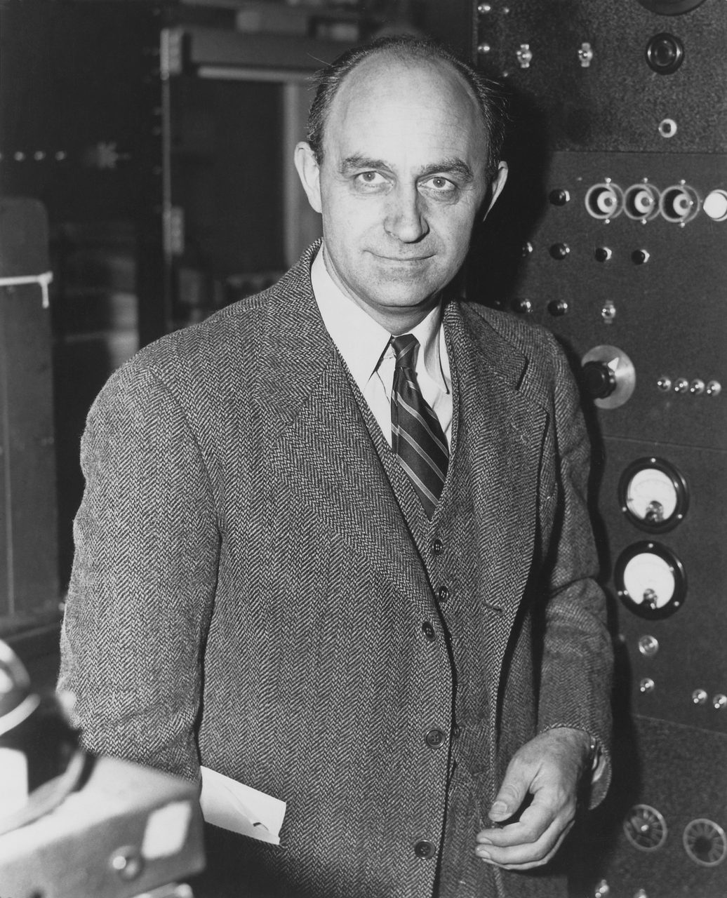 A black and white photograph of Enrico Fermi in a tweed jacket and tie, standing in front of a laboratory instrument panel.