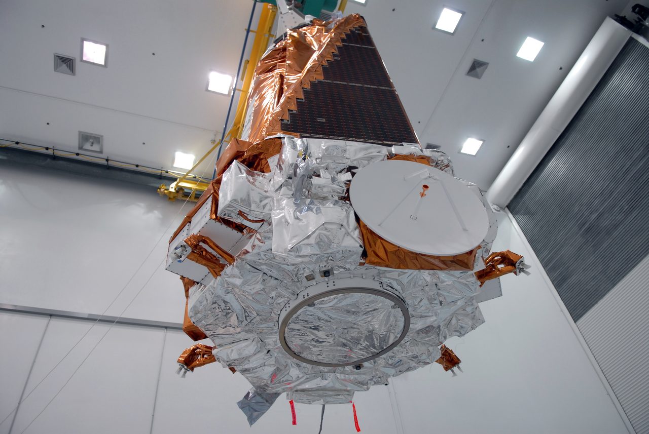 The Kepler spacecraft suspended in a cleanroom, wrapped in crinkled silver insulation foil, with a solar array wing above it.