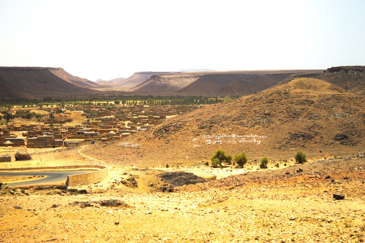 A settled oasis town in a valley on the Adrar Plateau of central Mauritania, with palm groves, mudbrick buildings, and a paved road set among rocky highland hills