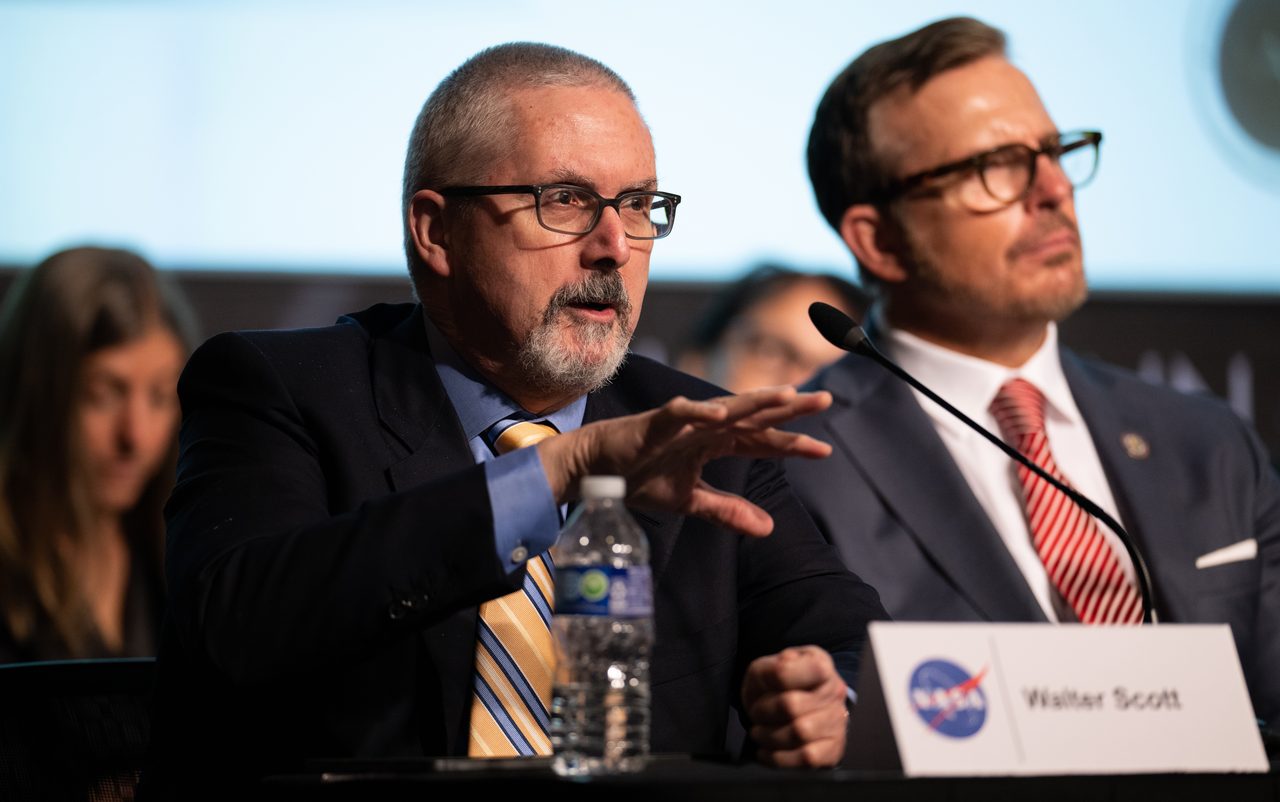 Two members of NASA's UAP Independent Study Team seated at a panel table during their public meeting, one speaking with a NASA nameplate reading Walter Scott visible on the desk