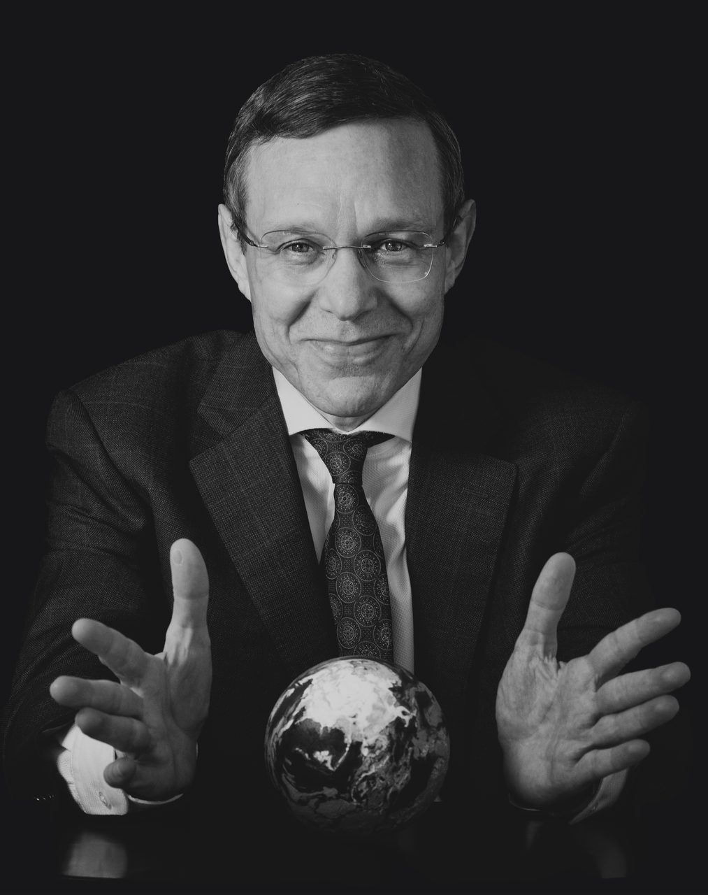 A black and white studio portrait of Avi Loeb in a dark suit, his open hands framing a small terrestrial globe on a dark table.