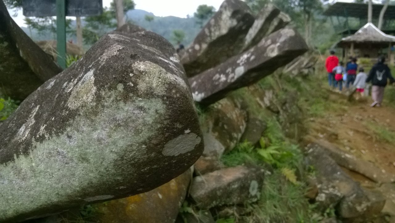 A close-up at Gunung Padang of a large rounded, lichen-mottled stone block filling the foreground, with tilted flat-faced slabs leaning together behind it, a dark signboard at the upper left and blurred visitors walking a path toward a pavilion at the right