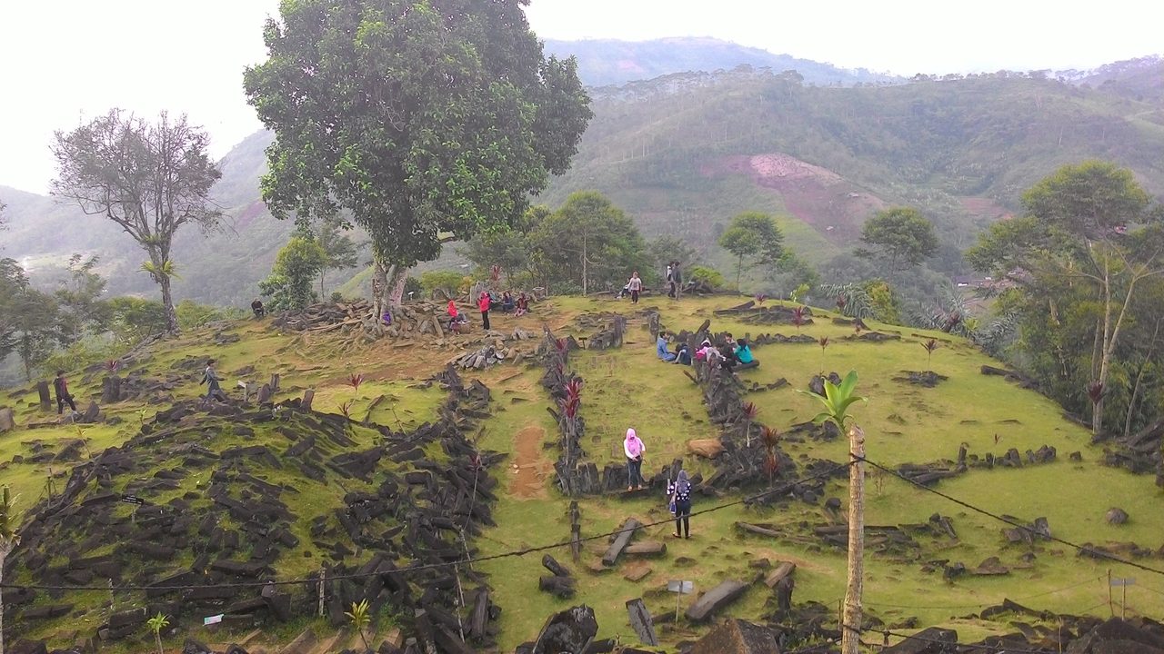 A wide elevated view across the topmost terrace of Gunung Padang, with a large spreading tree, visitors walking and sitting among low rows of columnar stones, open grass, and hazy green hills behind