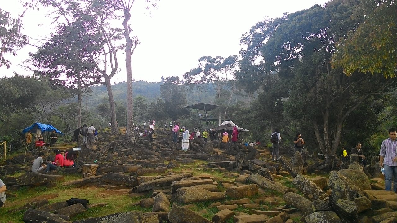 A crowd of visitors on the fourth terrace at Gunung Padang, with a metal-framed viewing tower at the center, a bright blue plastic tarpaulin over a stall at the left, a small open-sided hut at the right, dense fields of scattered columnar stone blocks in the foreground and a forested ridge behind