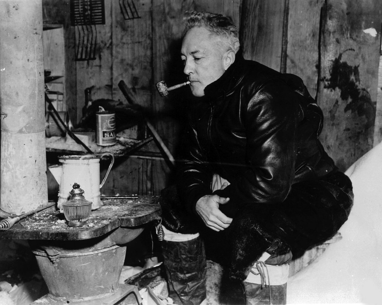 Rear Admiral Richard Byrd sitting inside his old Antarctic hut in 1947, a corncob pipe in his mouth, beside a stove with an oil lamp and an enamel pot on it