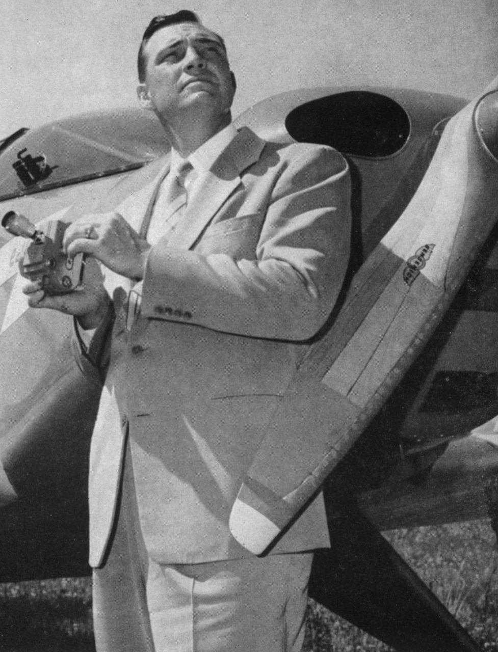 Black and white press portrait of Kenneth Arnold in a light suit standing beside a light aircraft, holding a small camera