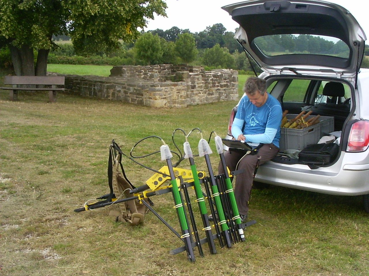 A modern archaeologist walking a pole-mounted magnetometer sensor across a field, transferring readings to a rugged laptop, at an archaeological site in Germany