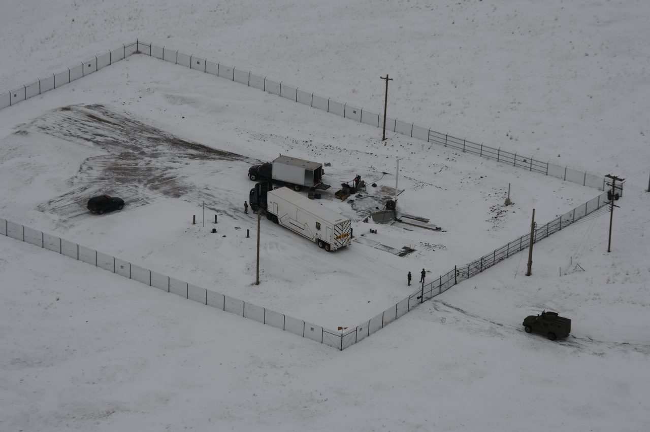 An aerial view of a snow-covered fenced launch facility with two trucks and personnel inside the perimeter
