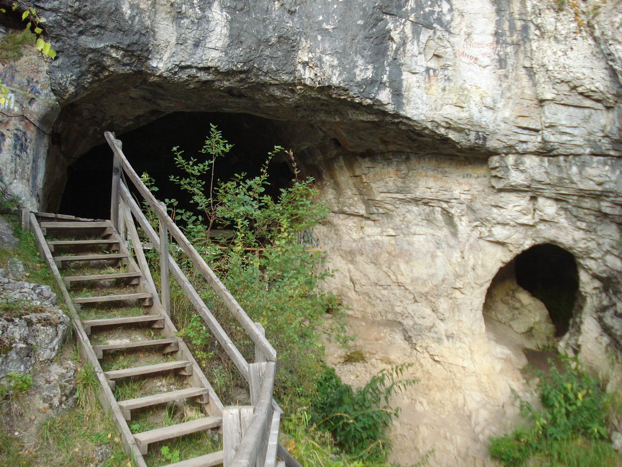 The entrance to Denisova Cave in the Altai region of Siberia: a wide dark cave mouth under an overhanging rock face, with a modern wooden staircase and handrail climbing to it