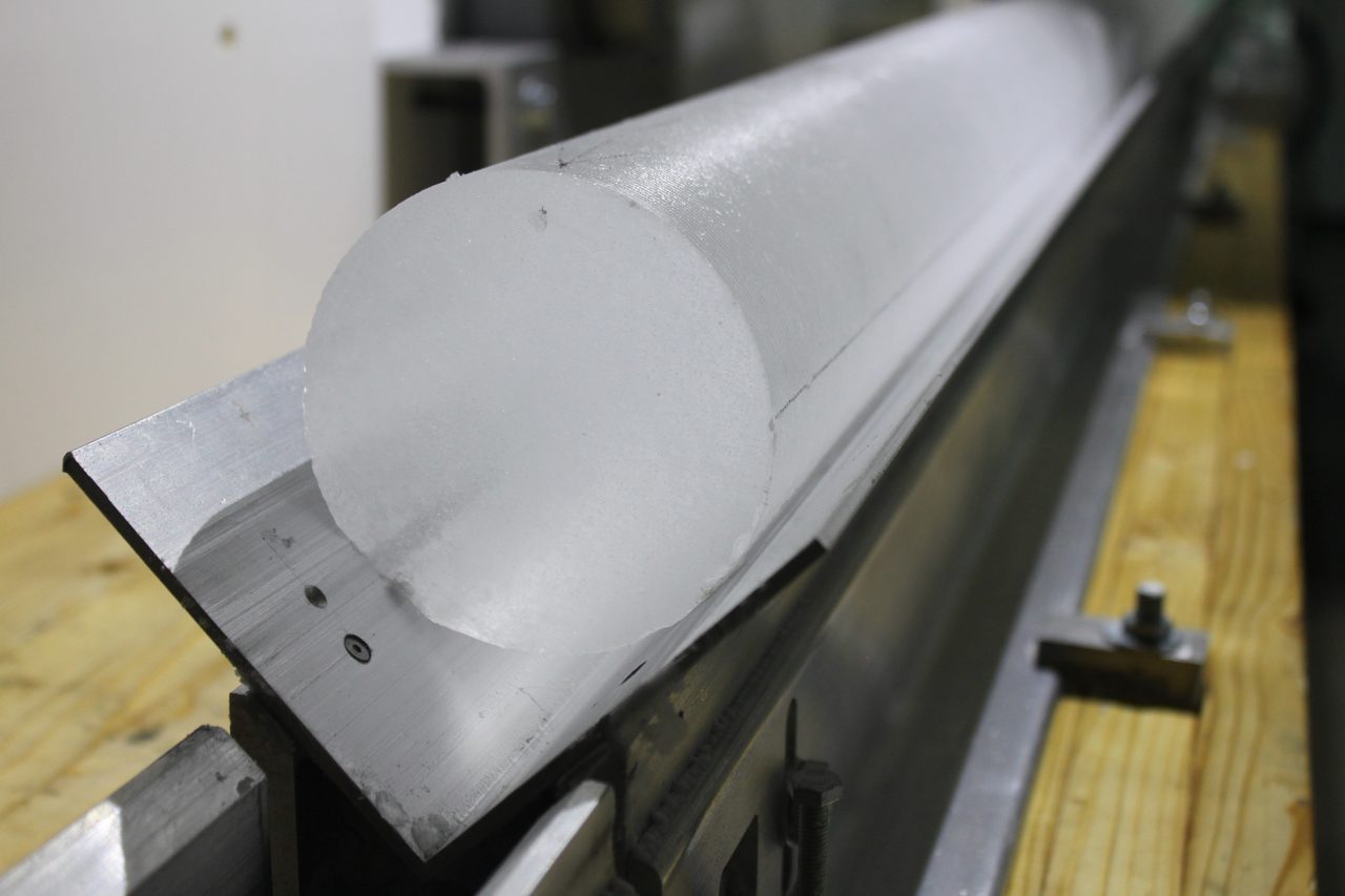 A freshly cut cylindrical section of the EastGRIP ice core resting on a light table, its cross-section showing faint horizontal layering in the clear ice.