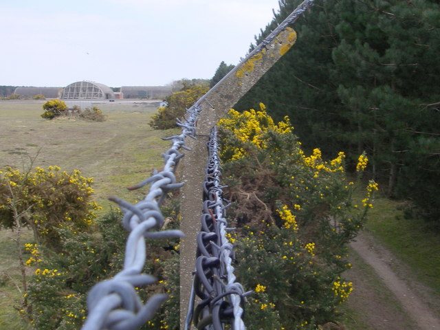 The perimeter fence of the former RAF Woodbridge airbase running through pine forest, with hardened aircraft shelters visible