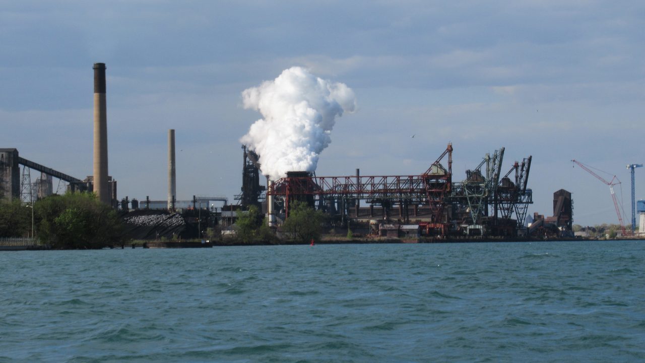 A wide daytime view of Zug Island, an industrial island in the Detroit River crowded with steel-mill structures, smokestacks, and storage tanks