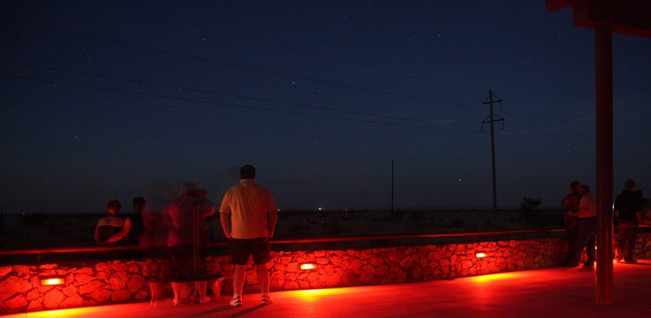 A long night exposure of visitors at the Marfa Lights viewing platform under a starry sky, with two small pale lights visible low on the desert horizon in the distance