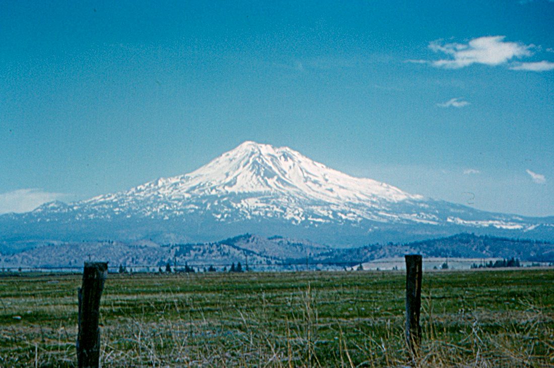 A snow-capped conical volcano rising above forested foothills under a clear blue sky
