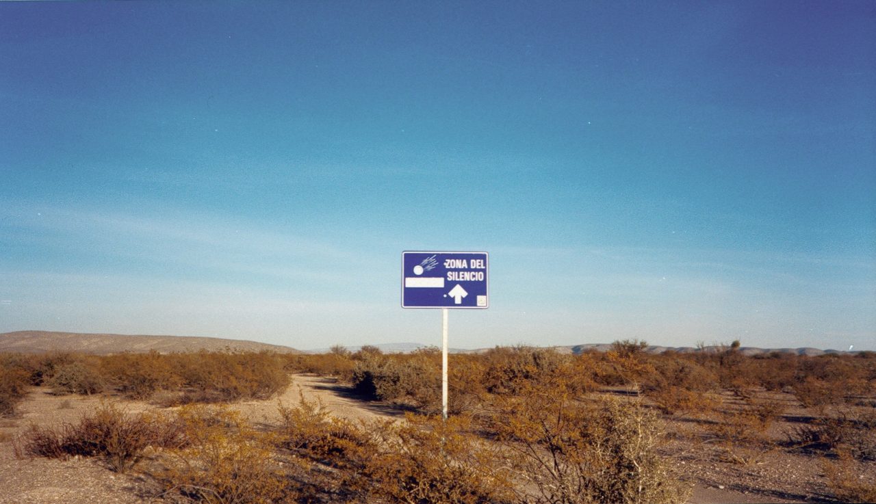 A blue roadside sign reading Zona del Silencio with a meteor icon, standing amid arid desert scrubland stretching to low hills in northern Mexico